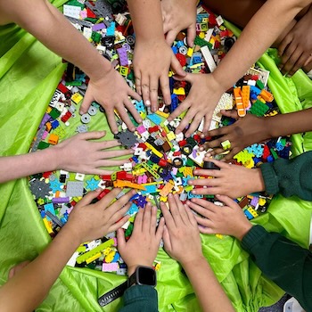 Children's hands reaching in from all sides to touch a large colourful pile of LEGO bricks spread on a bright green mat.